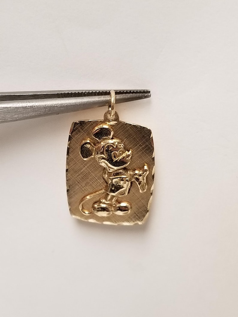 K&ouml;nnte beinhalten: Goldfarbener Anh&auml;nger mit einem erhabenen Bild von Mickey Mouse auf einer rechteckigen Platte. Der Anh&auml;nger wird von einer Pinzette gehalten. Die Platte hat eine strukturierte Oberfl&auml;che mit einem Rand.