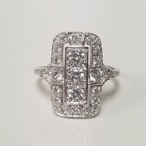 Op de afbeelding: Een zilveren ring met een rechthoekig ontwerp met een cluster van kleine diamanten en een grotere centrale diamant.