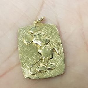 K&ouml;nnte beinhalten: Goldfarbener Anh&auml;nger mit rechteckiger, strukturierter Platte. Der Anh&auml;nger zeigt Mickey Mouse in klassischer Pose als Relief. Wahrscheinlich aus Gold gefertigt, als Schmuckst&uuml;ck zum Tragen gedacht.