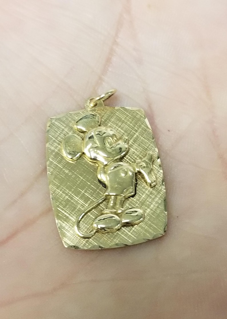 K&ouml;nnte beinhalten: Goldfarbener Anh&auml;nger mit rechteckiger, strukturierter Platte. Der Anh&auml;nger zeigt Mickey Mouse in klassischer Pose als Relief. Wahrscheinlich aus Gold gefertigt, als Schmuckst&uuml;ck zum Tragen gedacht.