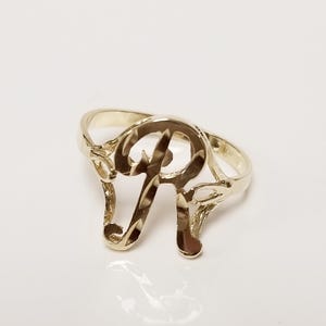 May include: A gold-colored ring featuring the letter "R" in a stylized, openwork design. The ring has a polished finish and is set against a white background. The ring is a piece of jewelry.