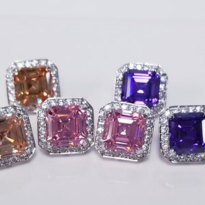 May include: Six square-shaped earrings with a halo of smaller stones surrounding a larger center stone. The center stones are in four colors: peach, pink, purple, and a darker purple.