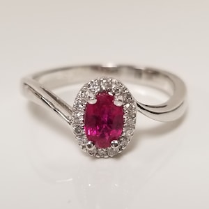 May include: A silver ring with a red oval ruby gemstone surrounded by a halo of smaller diamonds.