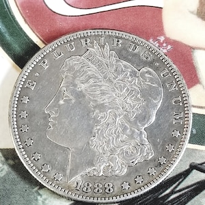 May include: A silver Morgan dollar coin from 1888, featuring a profile of a woman and the inscriptions "E PLURIBUS UNUM" and "LIBERTY". The coin has a detailed edge and is set against a colorful background.