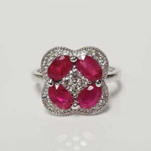 May include: A silver-toned ring featuring a clover-shaped setting. The center is adorned with four oval-cut deep red rubies arranged in a cross pattern, surrounded by small clear pavé-set stones and a milgrain edge on a polished metal band.