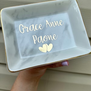 May include: A white marble trinket tray with a gold rim. The tray has the text "Grace Anne Paone" written on it in gold lettering. Two gold hearts are below the text.
