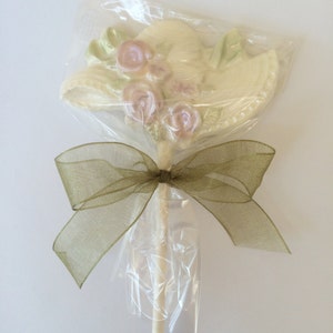 May include: A white chocolate hat with pink flowers on a stick, wrapped in clear cellophane and tied with a brown ribbon.