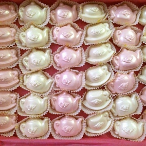 May include: A box of pink and white chocolate teapots. The teapots are arranged in rows and are all facing the same direction. The teapots are decorated with a delicate floral design.
