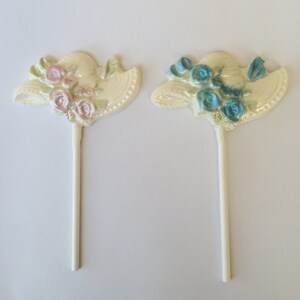May include: Two white chocolate lollipops shaped like hats with flowers. One hat has pink flowers and the other has blue flowers.