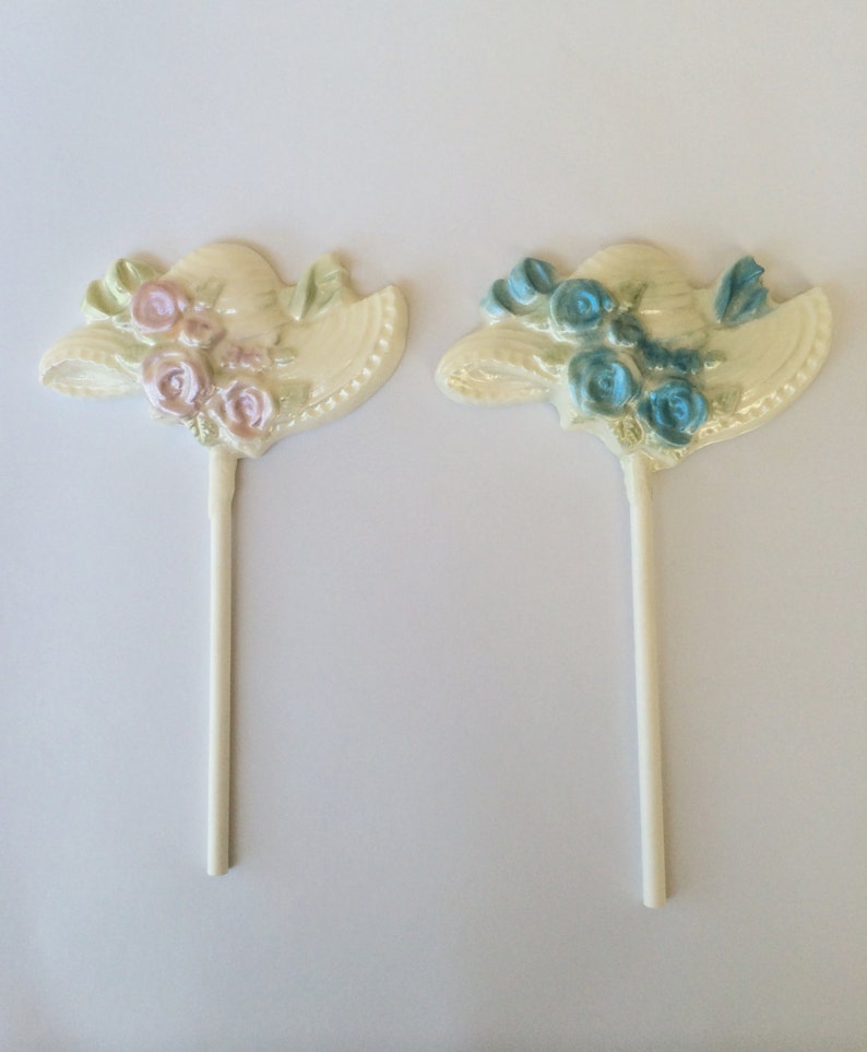 May include: Two white chocolate lollipops shaped like hats with flowers. One hat has pink flowers and the other has blue flowers.
