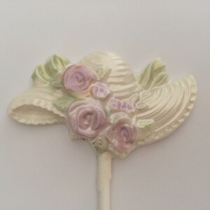 May include: A white ceramic hat with pink and green floral details. The hat is attached to a white stick.