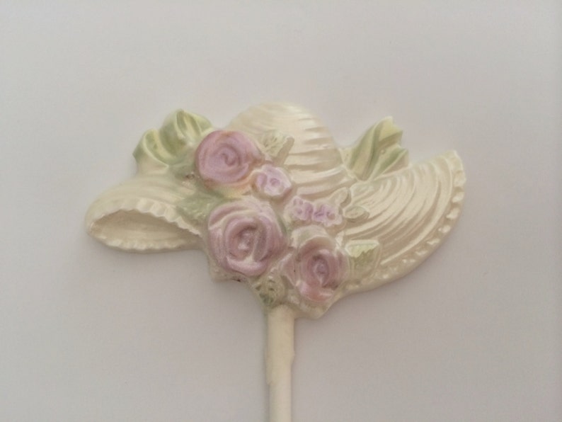 May include: A white ceramic hat with pink and green floral details. The hat is attached to a white stick.