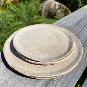 May include: A set of three ceramic plates in a light beige color. The plates are stacked on top of each other, with the smallest plate on top. The plates have a rough, textured surface.