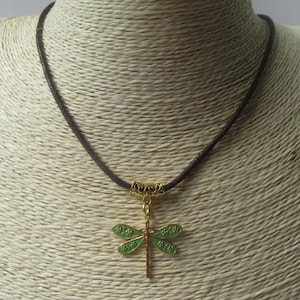 Peut inclure: Un collier en cordon marron avec un pendentif libellule doré. La libellule a des ailes en émail vert et un corps doré. Le collier est présenté sur un mannequin.
