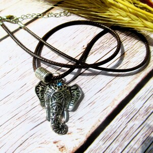 May include: A silver elephant pendant necklace with a brown cord. The elephant pendant features detailed patterns and a small blue gem. The necklace is presented on a light-coloured wooden surface.