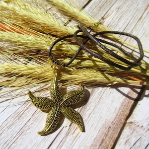 May include: A gold-toned starfish pendant necklace with a brown leather cord. The necklace is lying on a wooden surface with dried wheat stalks.