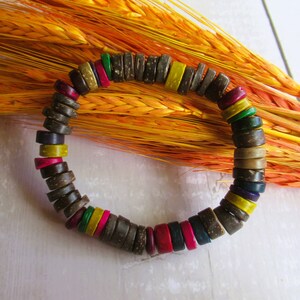May include: A wooden bead bracelet with dark brown, round beads and colourful, flat beads in yellow, pink, green, and red. The bracelet is set against a backdrop of orange wheat stalks.