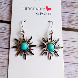 May include: Silver-coloured sunburst earrings with turquoise centres. The earrings are displayed on a white card with the text "Handmade with love" and a red heart. The background is a light-coloured wood grain.