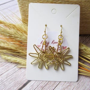 May include: Gold-tone earrings featuring a sun design. The earrings are displayed on a white card with the text "Ruby Rouge Jewelry" printed on it.