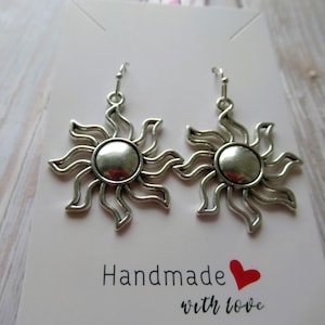 May include: Silver-coloured sun earrings, each with a central disc and radiating wavy lines. The earrings are presented on a white card with the words "Handmade with love" and a red heart.