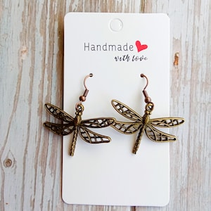May include: A pair of dragonfly earrings with a bronze finish. The earrings are displayed on a white card with the text "Handmade with love" and a red heart. The earrings have a detailed, open-work design.