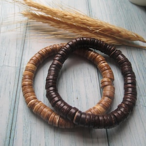 May include: Two wooden beaded bracelets in shades of brown and tan. The bracelets are made of small, round beads strung together. A sprig of wheat is in the background. The bracelets are on a light blue and white wooden surface.
