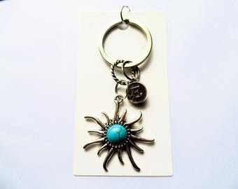 Türkiser Sonnen-Schlüsselanhänger und silberfarbener Initial-Charm an Karte mit Geschenkbeutel