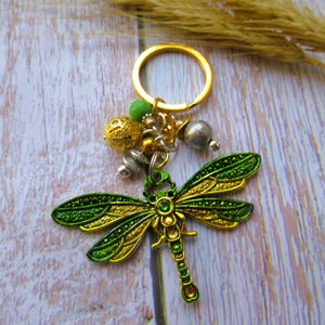 May include: A gold key chain with a green and gold dragonfly charm. The charm has intricate details and is accented with beads and other decorative elements.