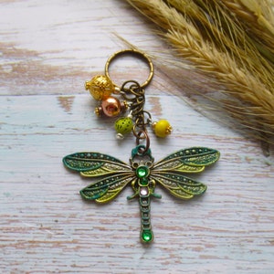 May include: A dragonfly keyring with a bronze-coloured ring and chain. The dragonfly has green and gold wings with a green body and jewel accents. The keyring also features decorative beads in gold, copper, and yellow.