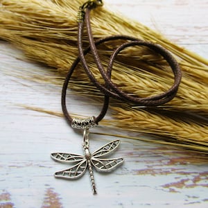 May include: A brown leather cord necklace with a silver dragonfly pendant. The necklace is lying on a bed of dried wheat stalks.