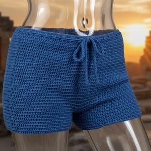 Könnte beinhalten: Blaue Häkelshorts mit Kordelzug in der Taille. Die Shorts sind einfarbig und scheinen handgefertigt zu sein. Der Hintergrund ist verschwommen, mit einem Sonnenuntergang und architektonischen Elementen.