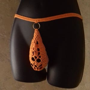 May include: Orange crochet thong with a decorative openwork design. The thong features a thin waistband and a teardrop-shaped front piece, connected by a gold-colored ring. The garment is displayed on a black mannequin.