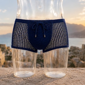 Puede incluir: Un bañador de color azul marino oscuro con un patrón de malla abierta estilo ganchillo en toda la prenda. El bañador incluye una cintura con cordón ajustable a juego con herretes metálicos en los extremos para un ajuste seguro.