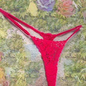 May include: A handmade, red crochet thong. The thong features a delicate, open weave design. The straps are thin and extend from the front to the back. The garment is displayed against a floral background.