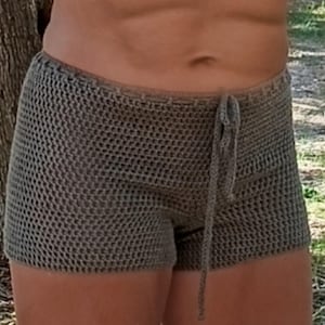 May include: Hand-crocheted shorts in a neutral gray color. The shorts feature a drawstring waist and a textured, open weave design. The garment is visible from the waist down, showcasing the intricate crochet work and casual style.