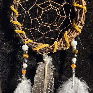 May include: A handcrafted dreamcatcher with a woven twig base, wrapped in yellow suede. The web is a geometric pattern. It features white, orange, and tiger-eye beads, and white and speckled feathers.