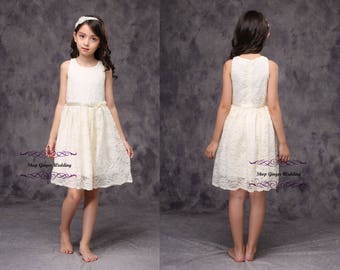 May include: A young girl wearing a white lace dress with a satin sash. The dress has a sleeveless design and a slightly flared skirt. The girl is standing on a gray background.