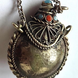Op de afbeelding: Een ronde, metalen hanger in de vorm van een kleine veldfles, met een getextureerd, verweerd zilverkleurig oppervlak en een gedraaide touwrand. De bovenkant is versierd met een waaiervormig filigraanelement, een oranje steen en twee kleine blauwe stenen.