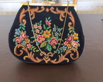 May include: A black handbag featuring a needlepoint floral design with pink, yellow, and blue flowers surrounded by tan scrollwork. The bag has a gold-tone metal frame, a matching gold-tone chain strap, and a top clasp closure mechanism.
