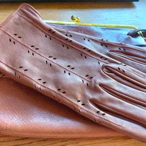 May include: A pair of brown leather gloves with decorative perforated leaf-like patterns on the back, resting on top of a matching brown leather clutch bag that features a gold-toned metal frame and a clasp closure at the top.