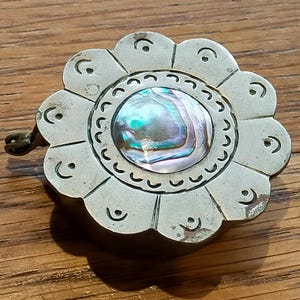 May include: A circular silver-toned metal pendant shaped like a flower with twelve petals. Each petal features a small engraved crescent moon symbol. The center is inlaid with a round, iridescent abalone shell piece showing green and purple hues.