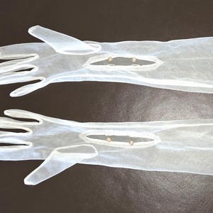 Peut inclure: Une paire de longs gants d'opéra blancs et transparents en tissu diaphane. Chaque gant présente une ouverture au niveau du poignet sur le dessus, fermée par deux petits boutons ronds en fausse perle crème, s'étendant sur l'avant-bras.