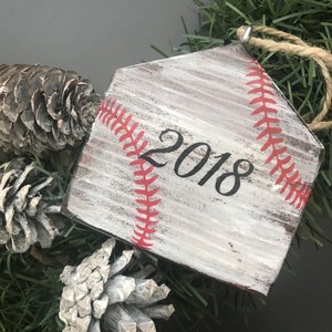 Pu&ograve; includere: Un ornamento in legno bianco a forma di home plate da baseball con cuciture rosse e l'anno 2018 scritto in nero. L'ornamento &egrave; appeso a una ghirlanda di pino artificiale verde.