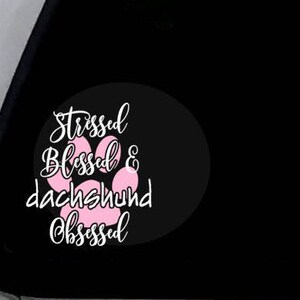 Op de afbeelding: Een zwarte en witte auto-raamsticker met een roze pootprint. De sticker zegt "Stressed Blessed & Dachshund Obsessed".