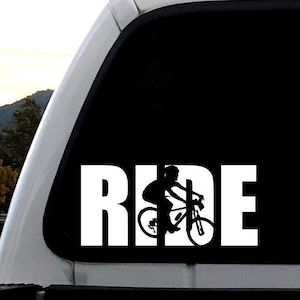 Puede incluir: Una silueta en blanco y negro de una persona montando en bicicleta con la palabra "RIDE" en letras blancas sobre un fondo negro. La imagen está en la ventana trasera de una camioneta blanca.