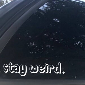 K&ouml;nnte beinhalten: Eine schwarze Autot&uuml;r mit einem wei&szlig;en Vinyl-Aufkleber, auf dem "stay weird." steht.