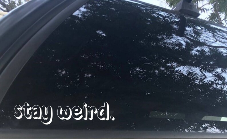 K&ouml;nnte beinhalten: Eine schwarze Autot&uuml;r mit einem wei&szlig;en Vinyl-Aufkleber, auf dem "stay weird." steht.