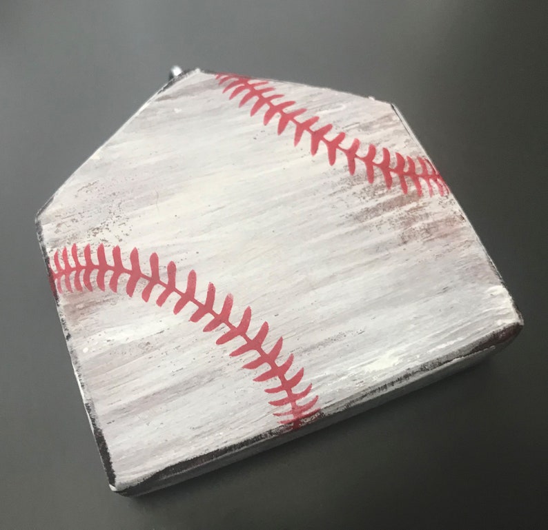 Pu&ograve; includere: Un piatto di casa in legno bianco con cuciture da baseball rosse dipinte sulla superficie.