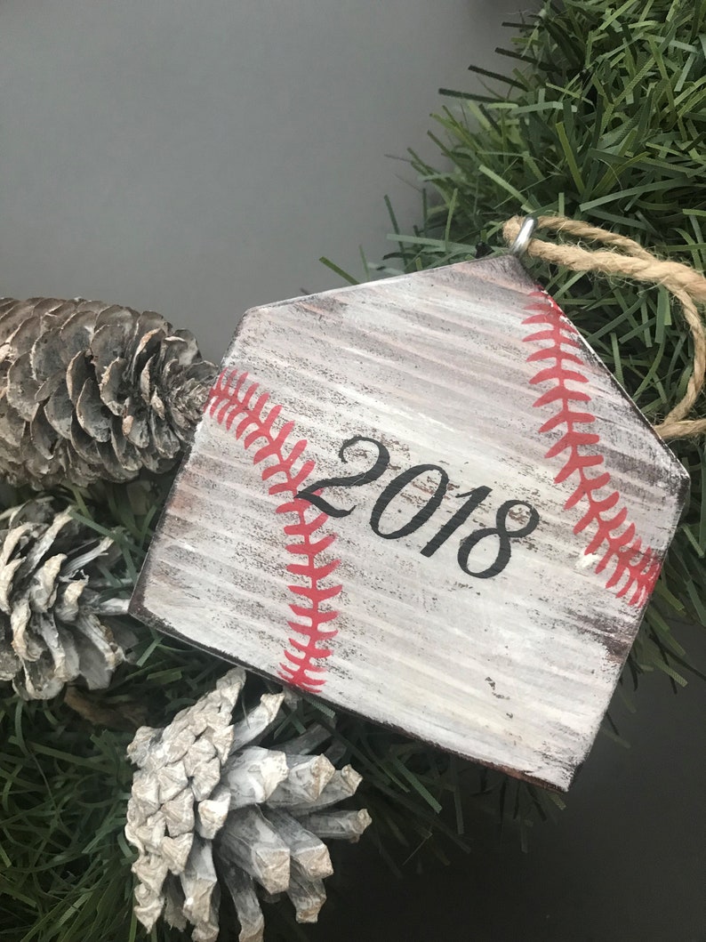 Pu&ograve; includere: Un ornamento in legno bianco a forma di home plate da baseball con cuciture rosse e l'anno 2018 scritto in nero. L'ornamento &egrave; appeso a una ghirlanda di pino artificiale verde.