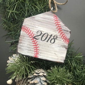 Pu&ograve; includere: Un ornamento di Natale in legno bianco a forma di baseball con cuciture rosse e l'anno 2018 dipinto su di esso.